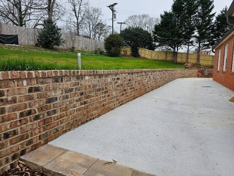 Brick retaining wall beside a long concrete driveway next to a brick building and grassy slope.