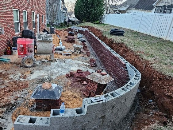Curved concrete retaining wall under construction beside a brick house and white fence in a backyard