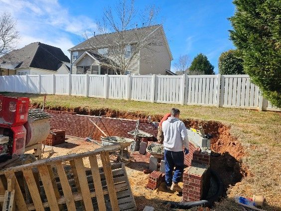 Workers in a backyard excavation beside a red excavator and wooden retaining wall