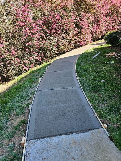Curved concrete sidewalk beside pink flowering shrubs and green grass on a sunny hillside