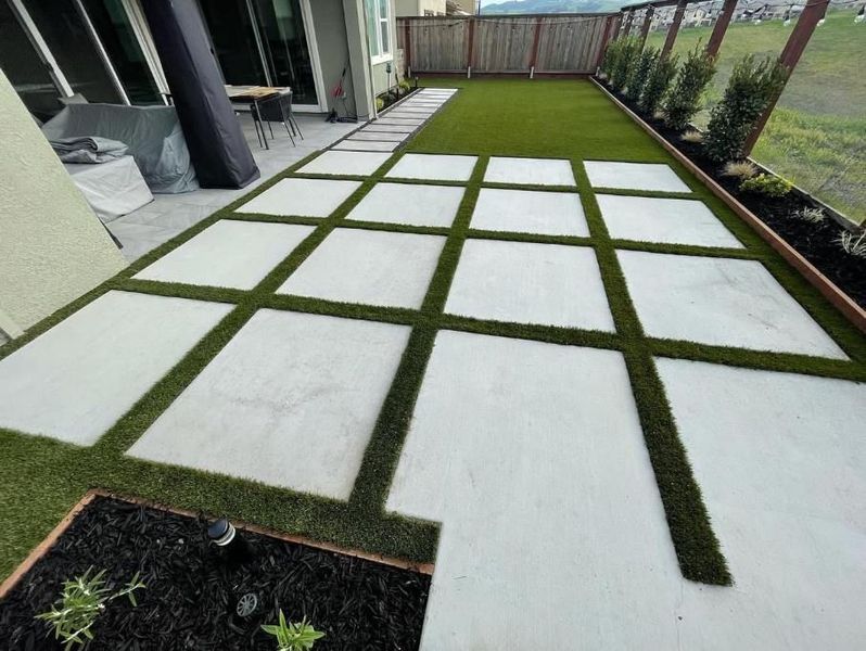 Backyard patio with large white pavers set in green grass, beside a house and fence