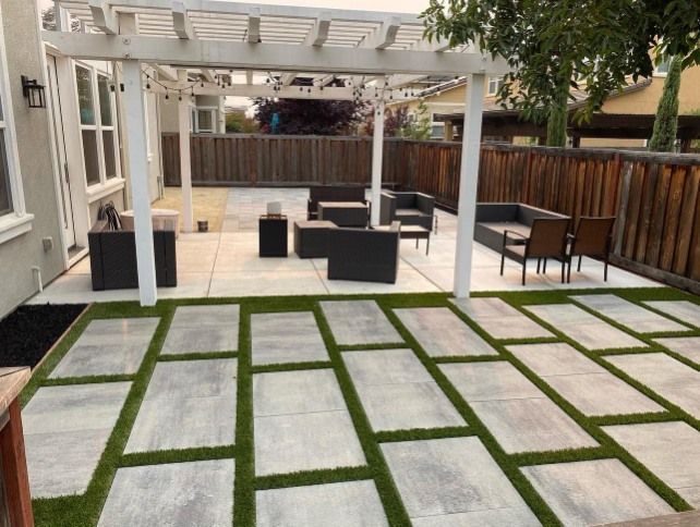Backyard patio with white pergola, gray seating, wooden fence, and stone pavers in grass