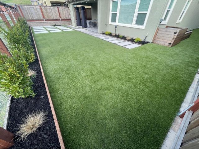 Small fenced backyard with artificial grass, stepping stones, and a patio beside a house.