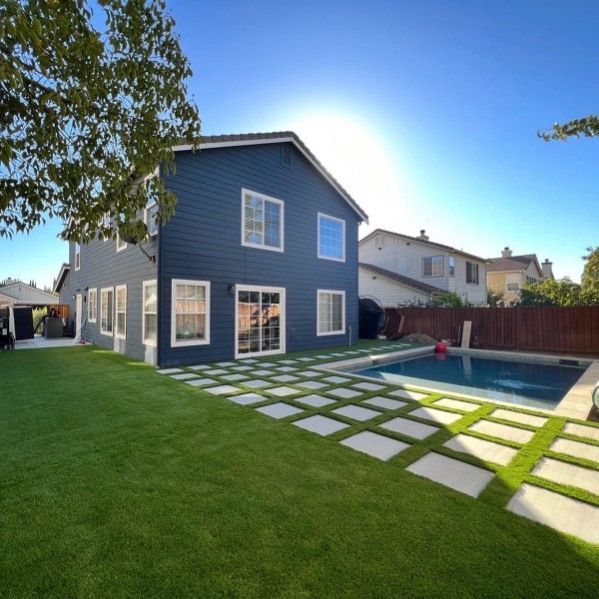 Backyard with blue two-story house, lawn, stepping stones, and a swimming pool under bright sun