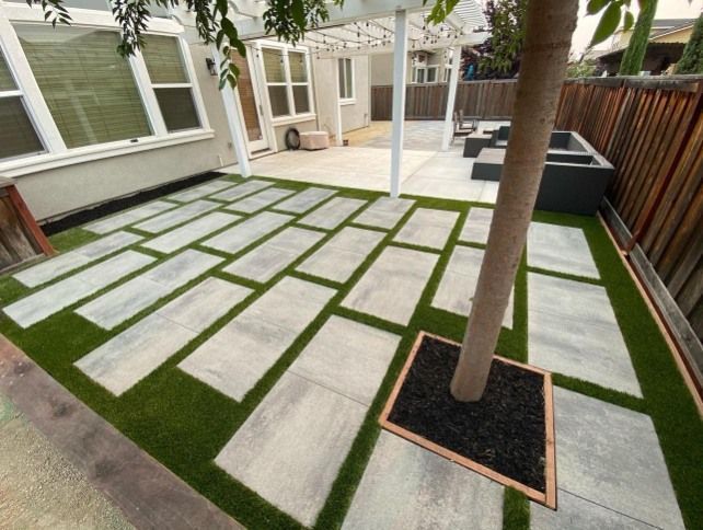 Modern backyard patio with large white stepping stones, green turf, and a tree in a square planter