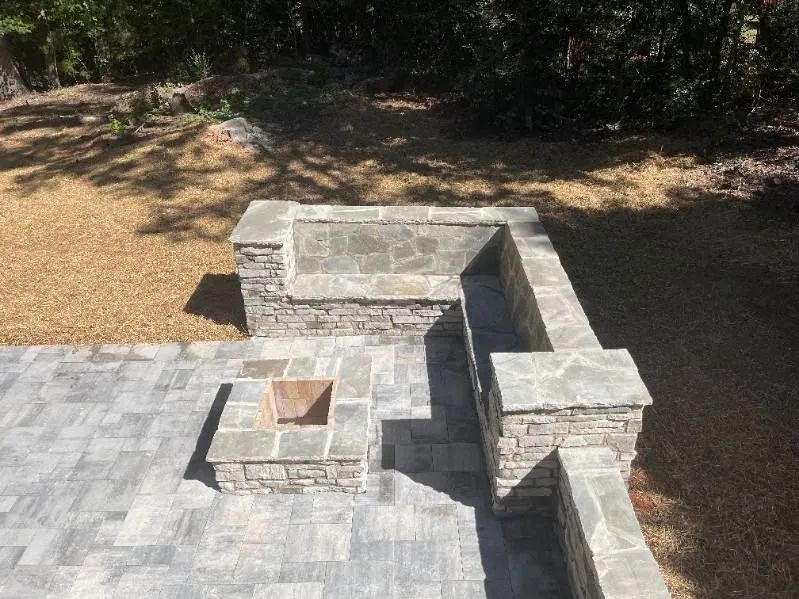 Stone outdoor patio with built-in benches and a square fire pit in a sunny yard