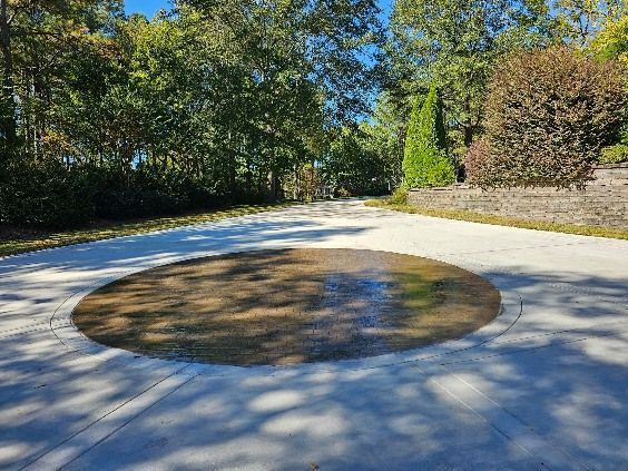 Circular stain on a paved driveway, surrounded by trees and stone walls in sunlight.