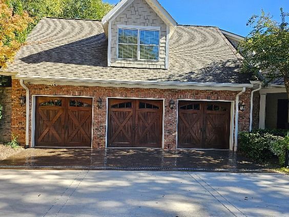 Stone house with three wooden garage doors and a dormer window, shaded by trees.