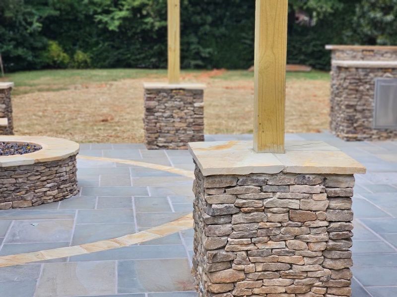 Stone patio with wooden posts and a circular stone fire pit in a landscaped yard