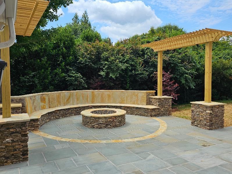 Stone patio courtyard with circular fire pit, stone benches, and wooden pergolas under a partly cloudy sky