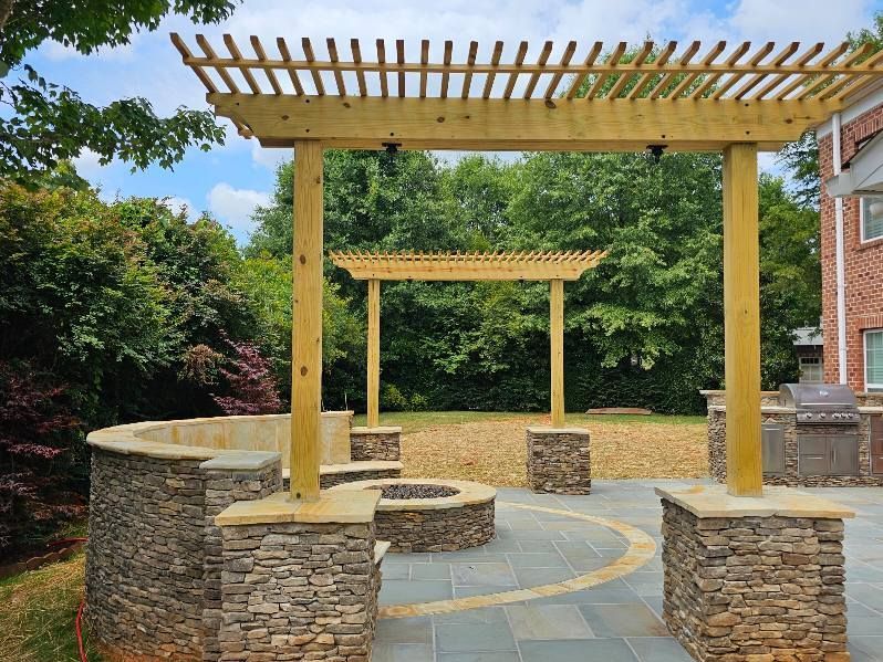 Stone patio with curved seating, wooden pergola, and garden greenery beside a brick house