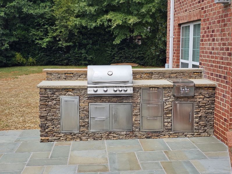 Outdoor built-in stainless steel grill and cabinets on a stone patio beside a brick house