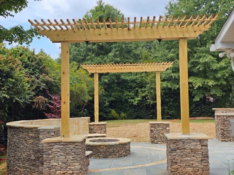 Wooden pergolas and stone seating in a landscaped outdoor patio with trees in the background