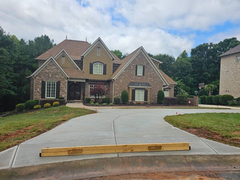 Large brick suburban house with a wide driveway and landscaped front yard under a cloudy sky