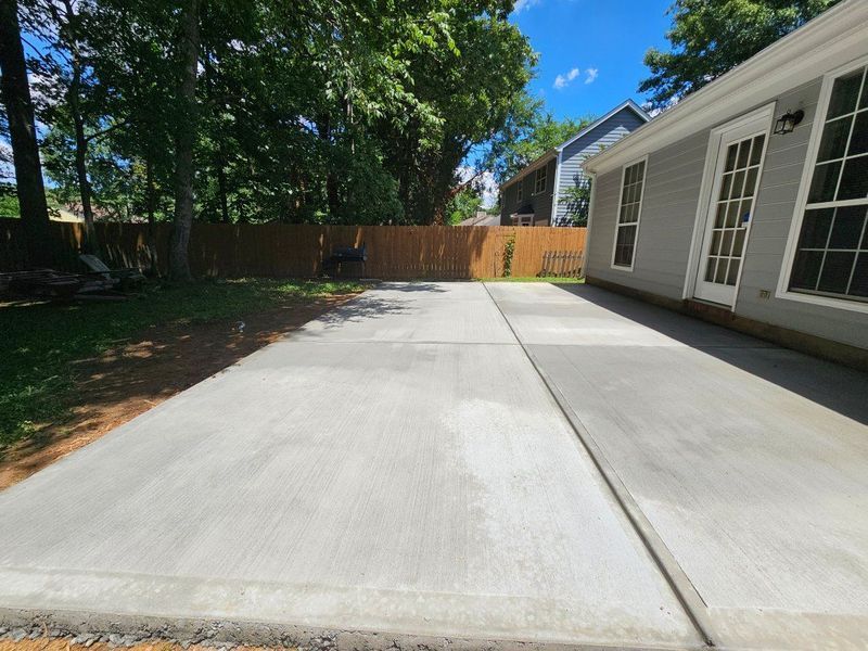 Concrete backyard patio beside a white house, with trees and a wooden fence in the background