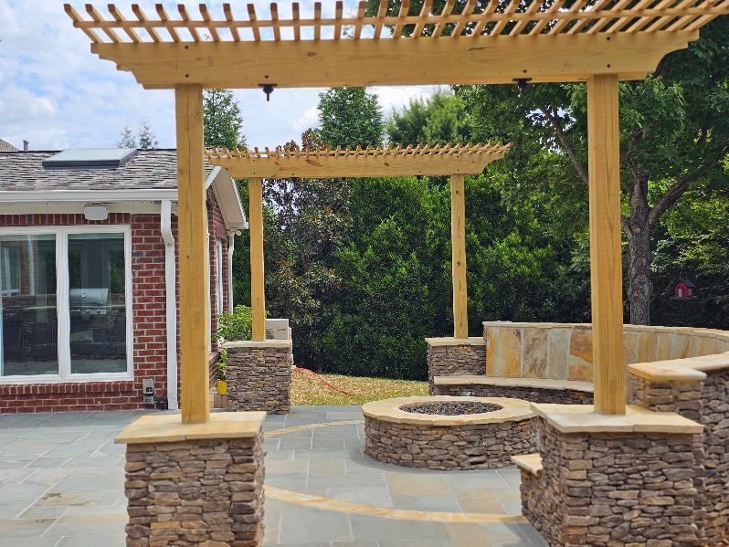Wooden pergola over a stone patio with built-in seating and a circular fire pit in a backyard.