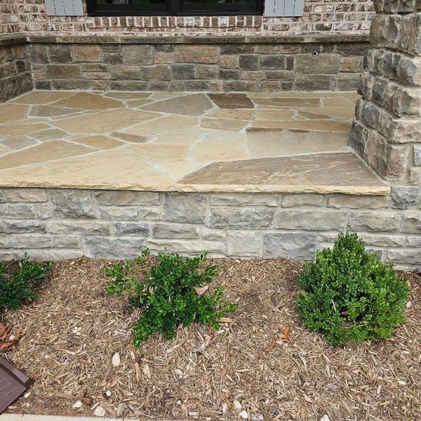 Stone front porch with a raised ledge and three small shrubs in a mulched flower bed