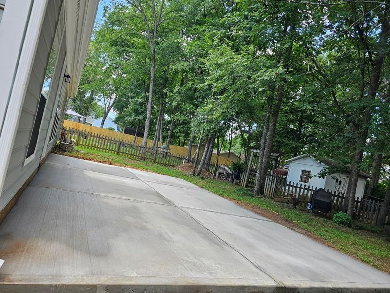 Concrete driveway beside a house, leading to a fenced, tree-filled backyard on an overcast day