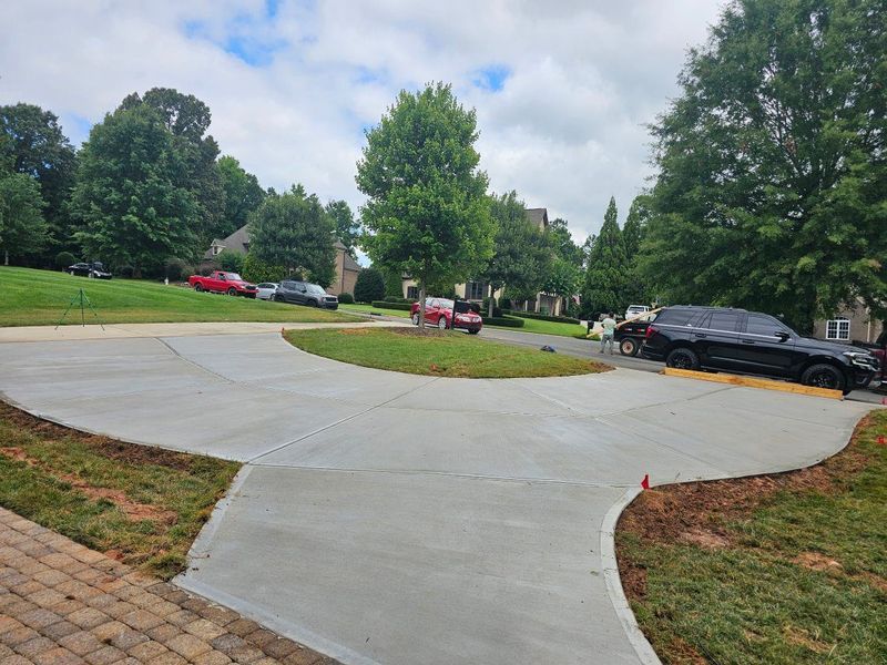 Fresh concrete driveway curves through a suburban yard with parked cars and trees under a cloudy sky