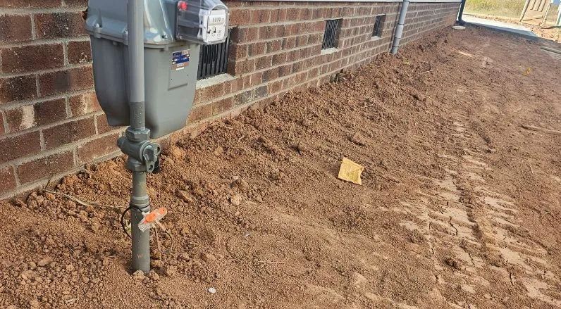 Gas meter and regulator mounted on a brick house wall beside freshly dug soil along the foundation.