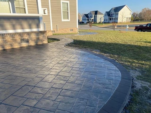 Curved stamped concrete patio beside a tan house, bordered by grass in a suburban neighborhood.