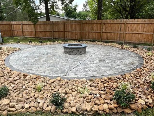 Backyard patio with a circular stone fire pit, surrounded by a rock border and wooden fence