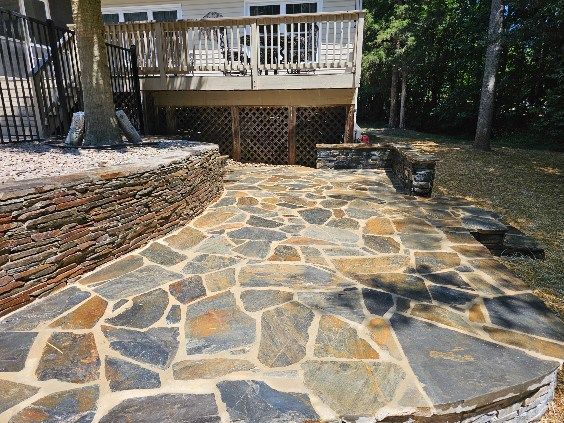 Stone patio and walkway leading to a wooden deck in a wooded backyard, bordered by stacked stone walls.