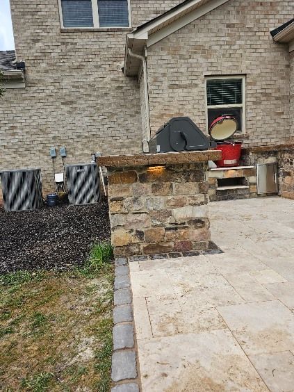 Stone outdoor grill beside a stone patio and brick house wall, with a covered grill and red bucket nearby