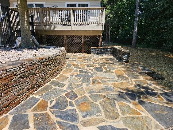 Stone patio leading to a wooden deck beside a house, with stacked stone retaining walls and trees