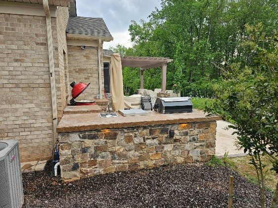 Patio with stone retaining wall, grill, red chair, and covered seating beside a brick house and trees