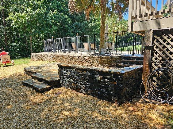 Stone patio steps beside a low wall and deck, with a coiled hose and trees in a sunny backyard