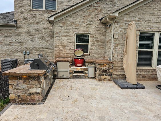Stone patio with built-in grill, fire pit, and covered umbrella beside a brick house.