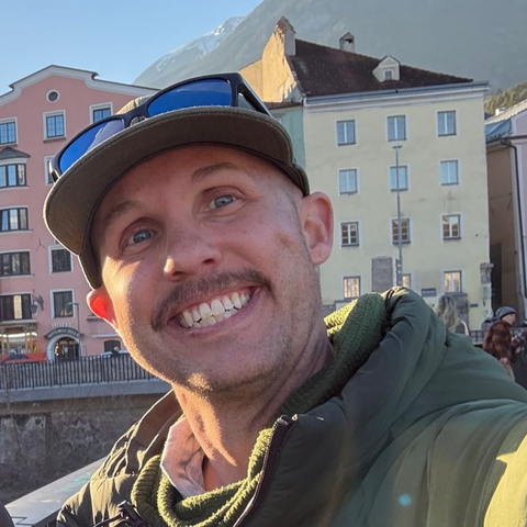 Man smiles, wearing a cap and sunglasses, in front of colorful buildings and mountains.