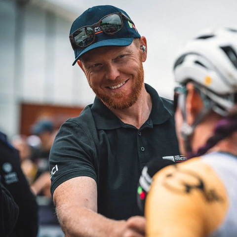 Man in cap smiles, shaking hands with cyclist in helmet and jersey at an outdoor event.