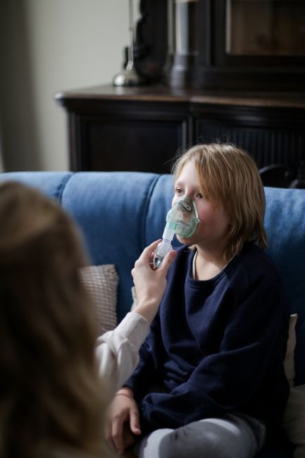 Un niño recibe tratamiento con nebulizador en un sofá azul, mientras un adulto le sostiene la mascarilla.