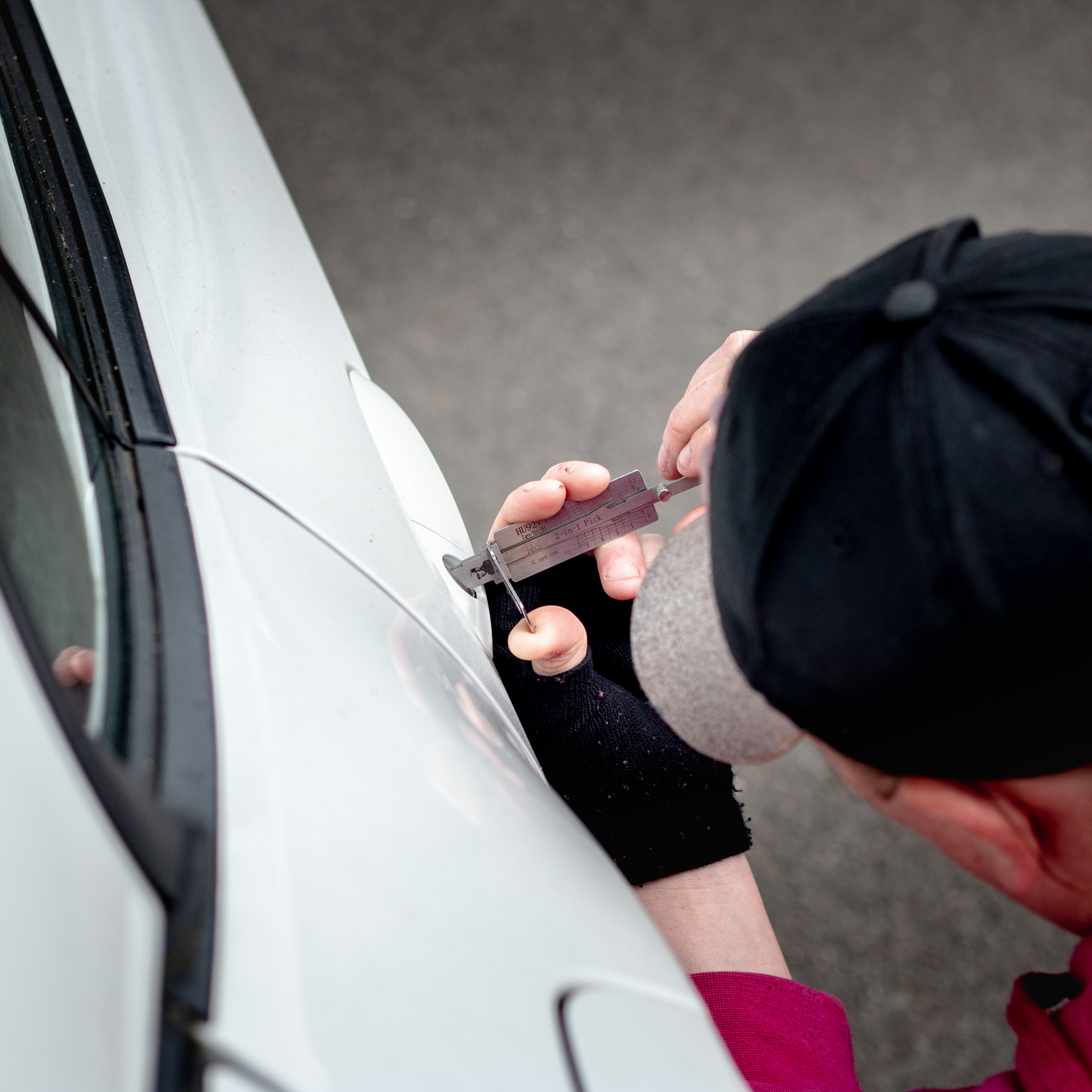 Person using lock-picking tools on a white car door, wearing a black cap and glove.