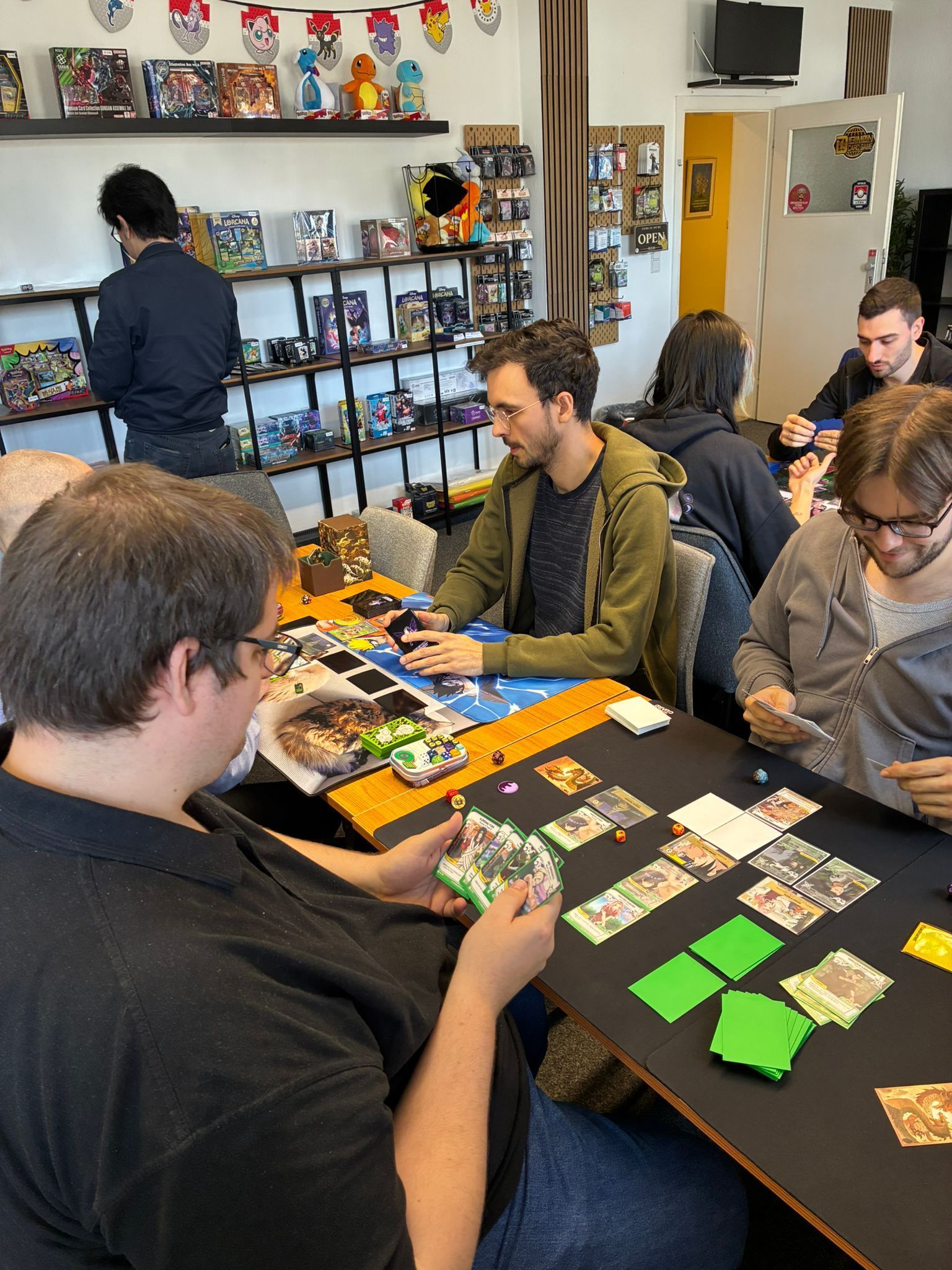 People playing a colorful board game at a table in a game store, with shelves of games in the background.
