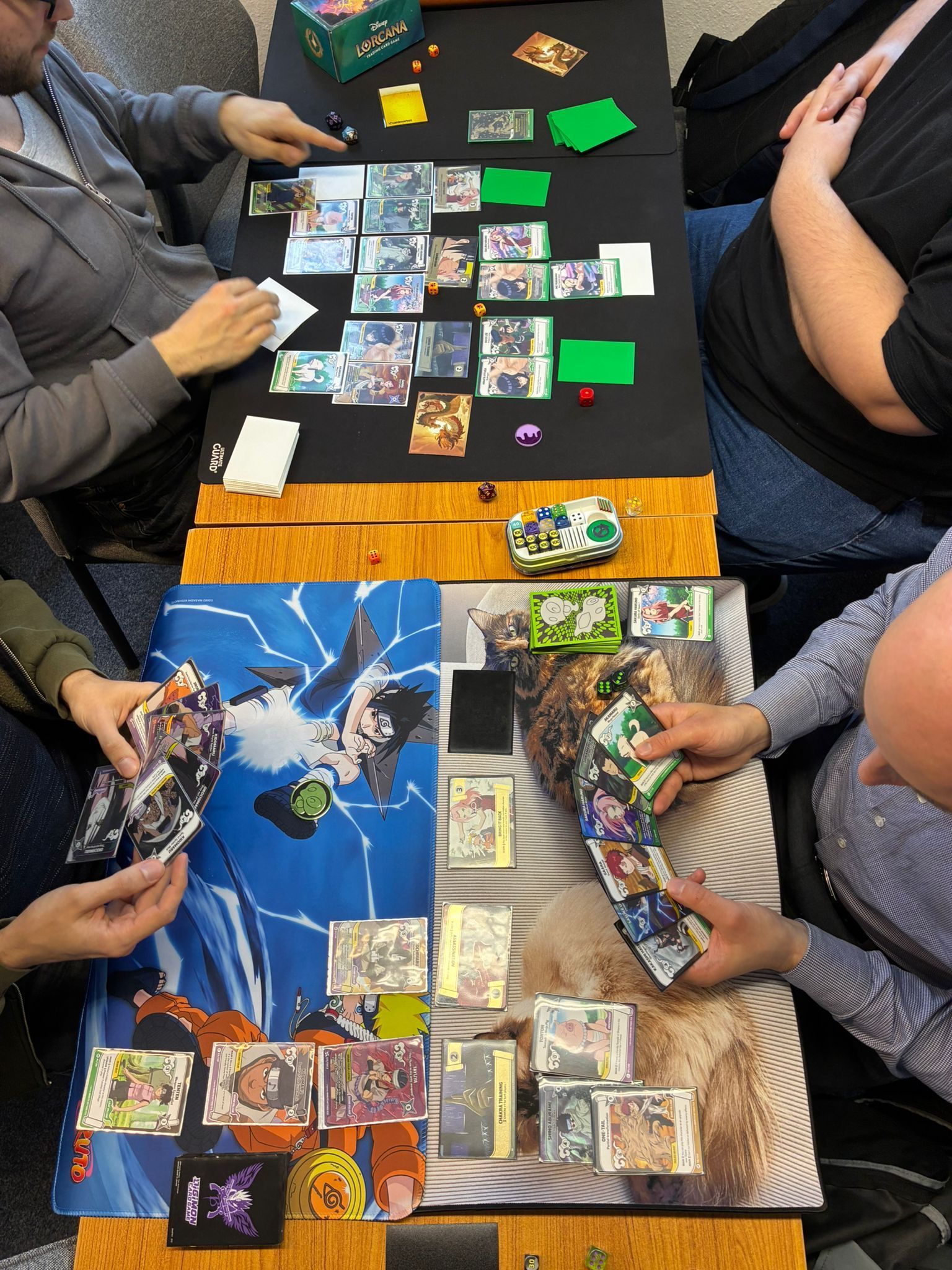 Three people play a card game on a table covered with colorful cards and playmats.