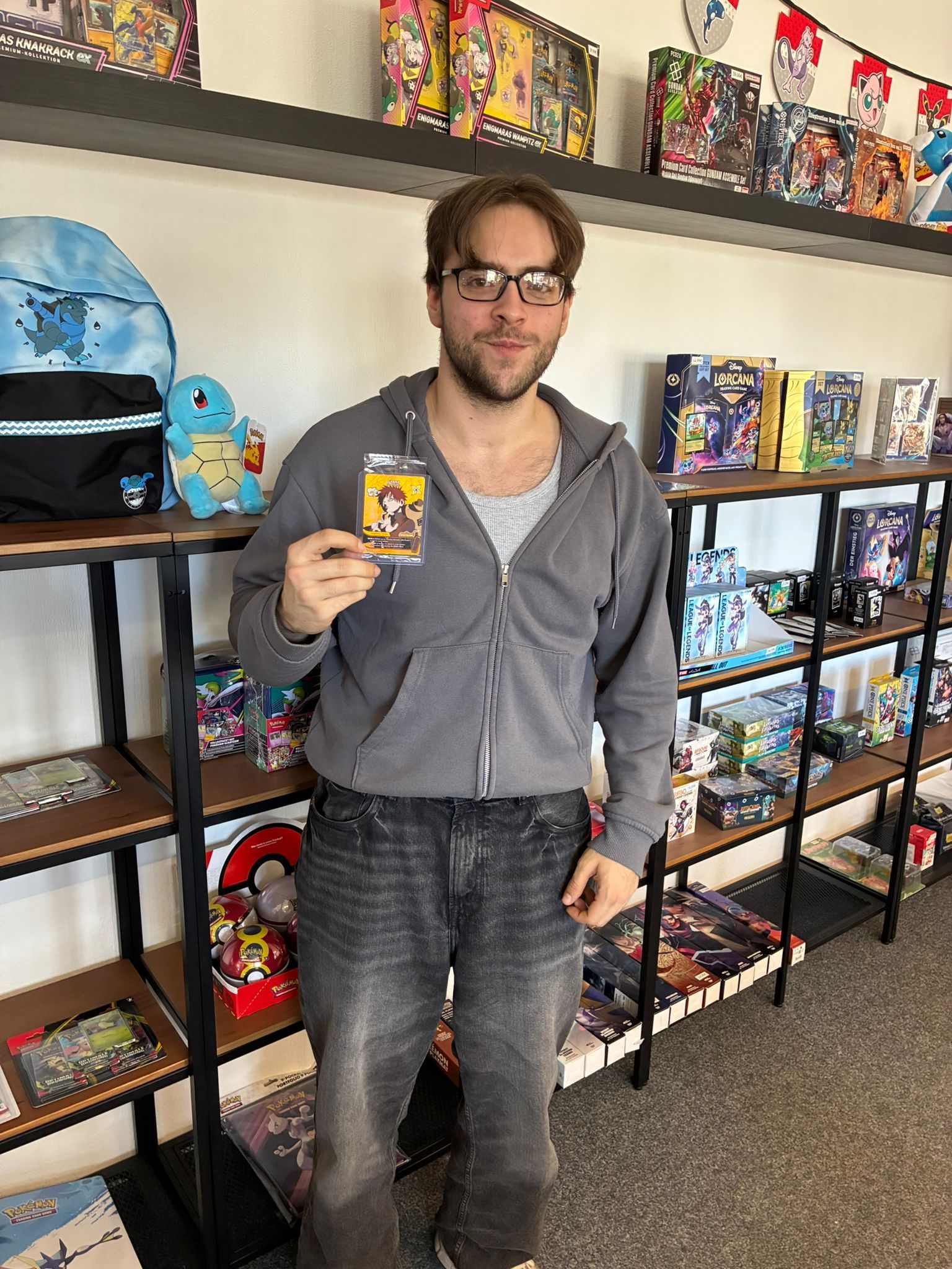 Man holding a trading card in front of shelves filled with collectible card boxes and toys