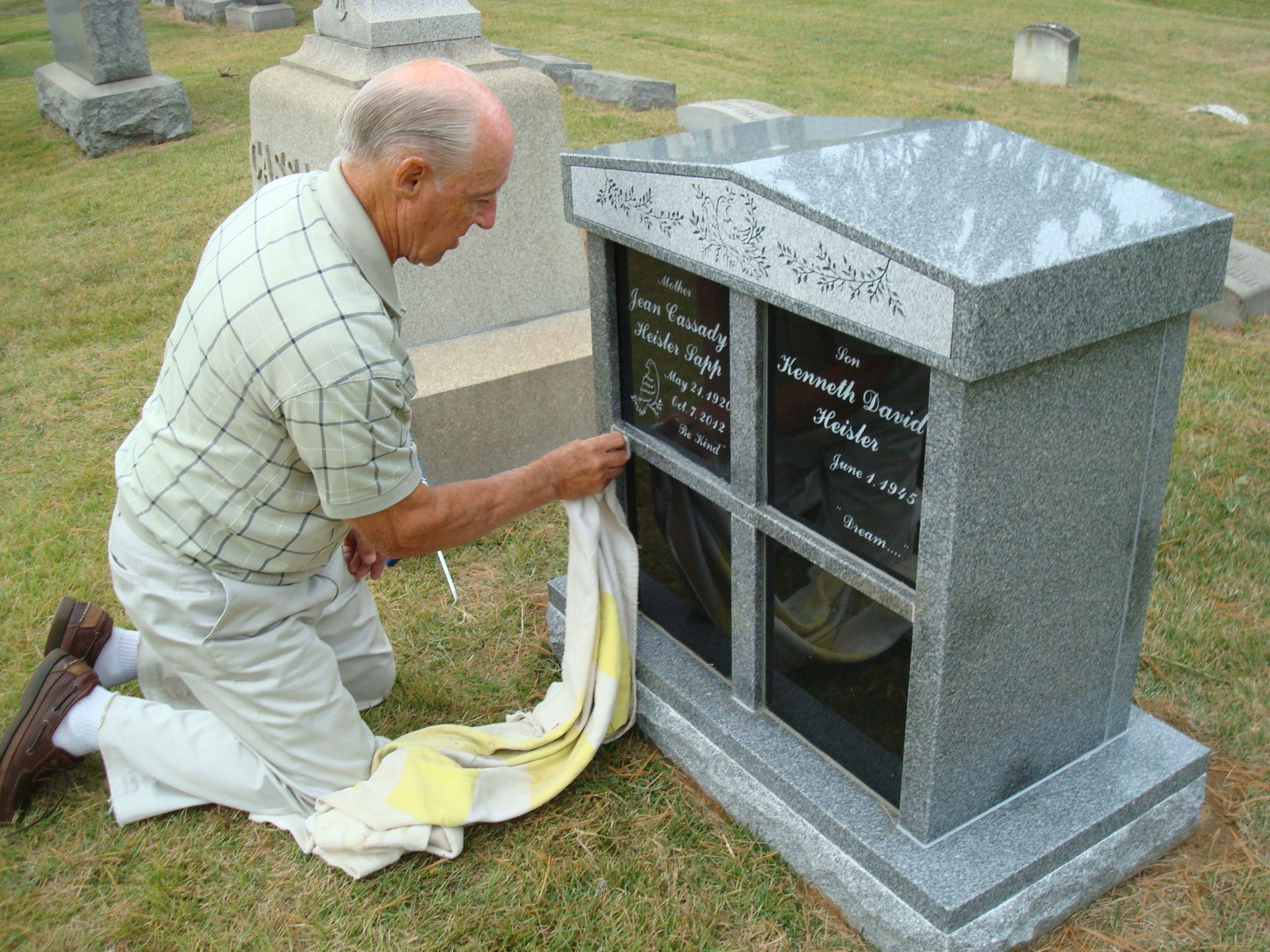 An older person kneels on grass, cleaning a polished gray granite monument in a cemetery with a white cloth.