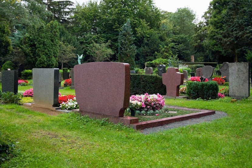 A cemetery with various gravestones and colorful flower beds arranged on manicured green grass under leafy trees.