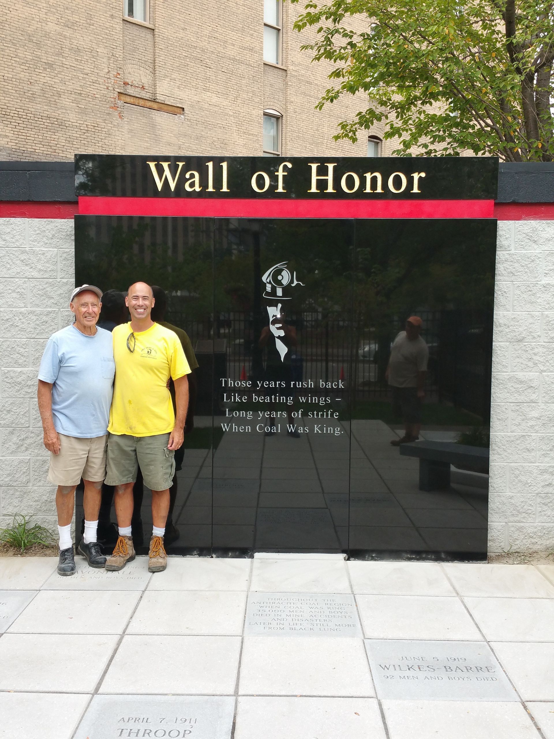 Two people stand side-by-side smiling in front of a black