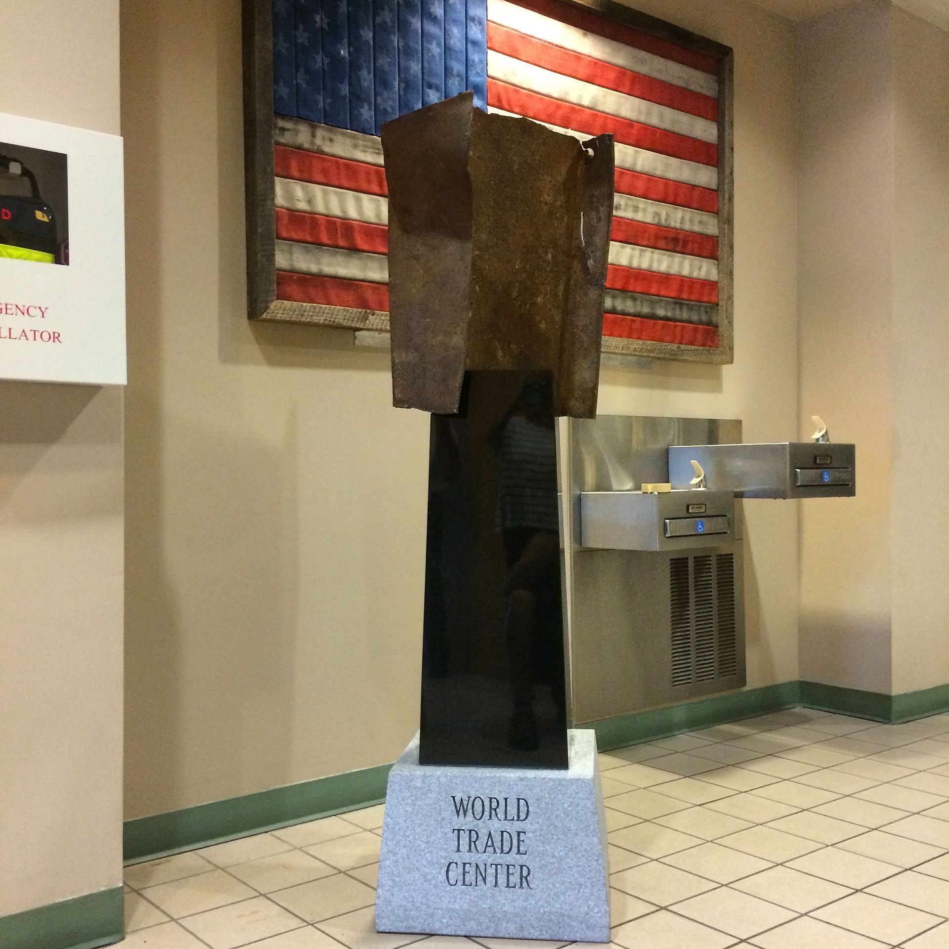 A World Trade Center memorial display featuring a steel beam fragment on a pedestal before a tattered American flag.
