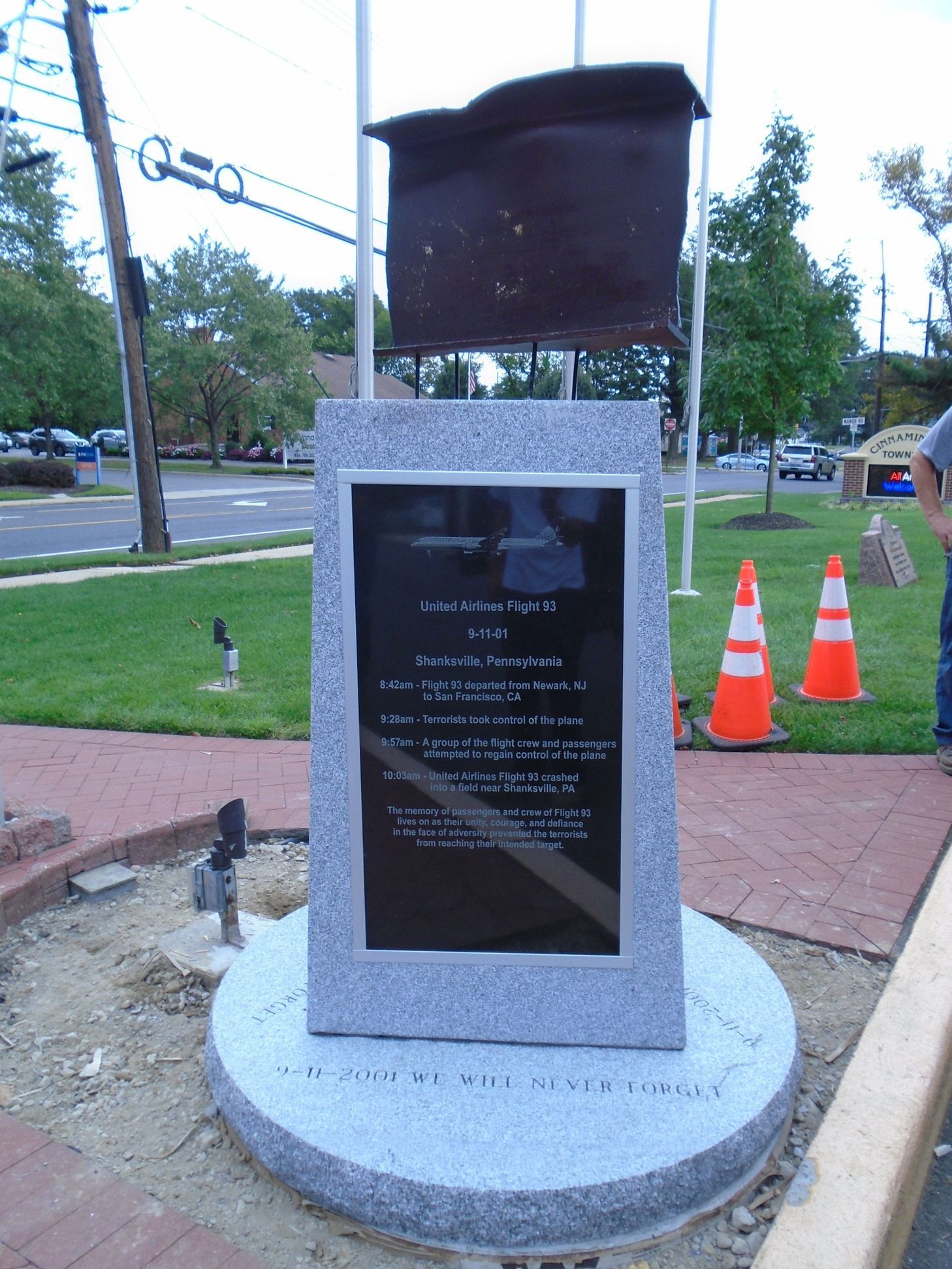 A memorial monument with a rusted metal beam atop a granite base, standing on a sidewalk near a grassy field and road.