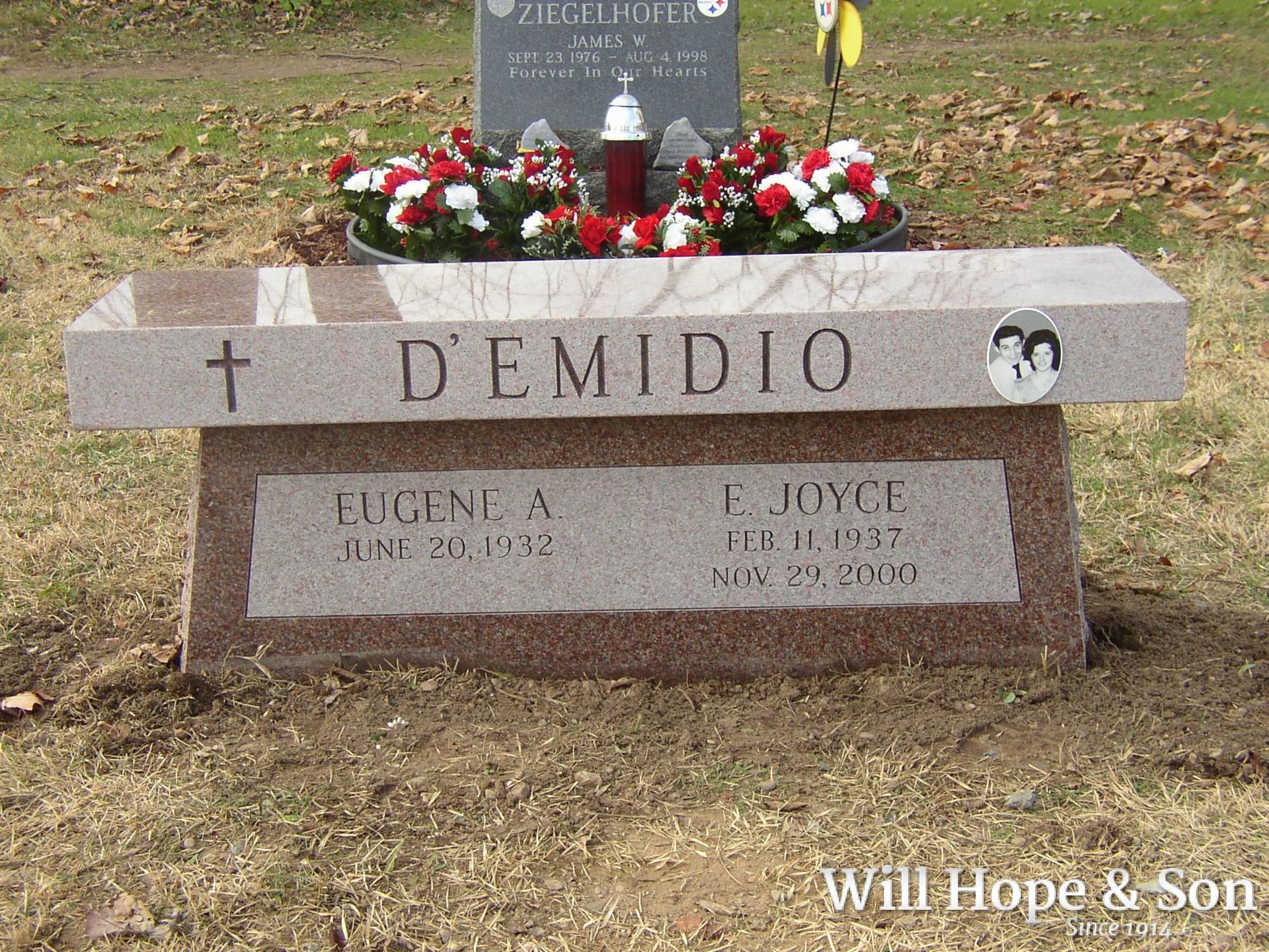 A granite bench headstone for the D’Emidio family, inscribed with names and dates, situated on a grassy cemetery plot.