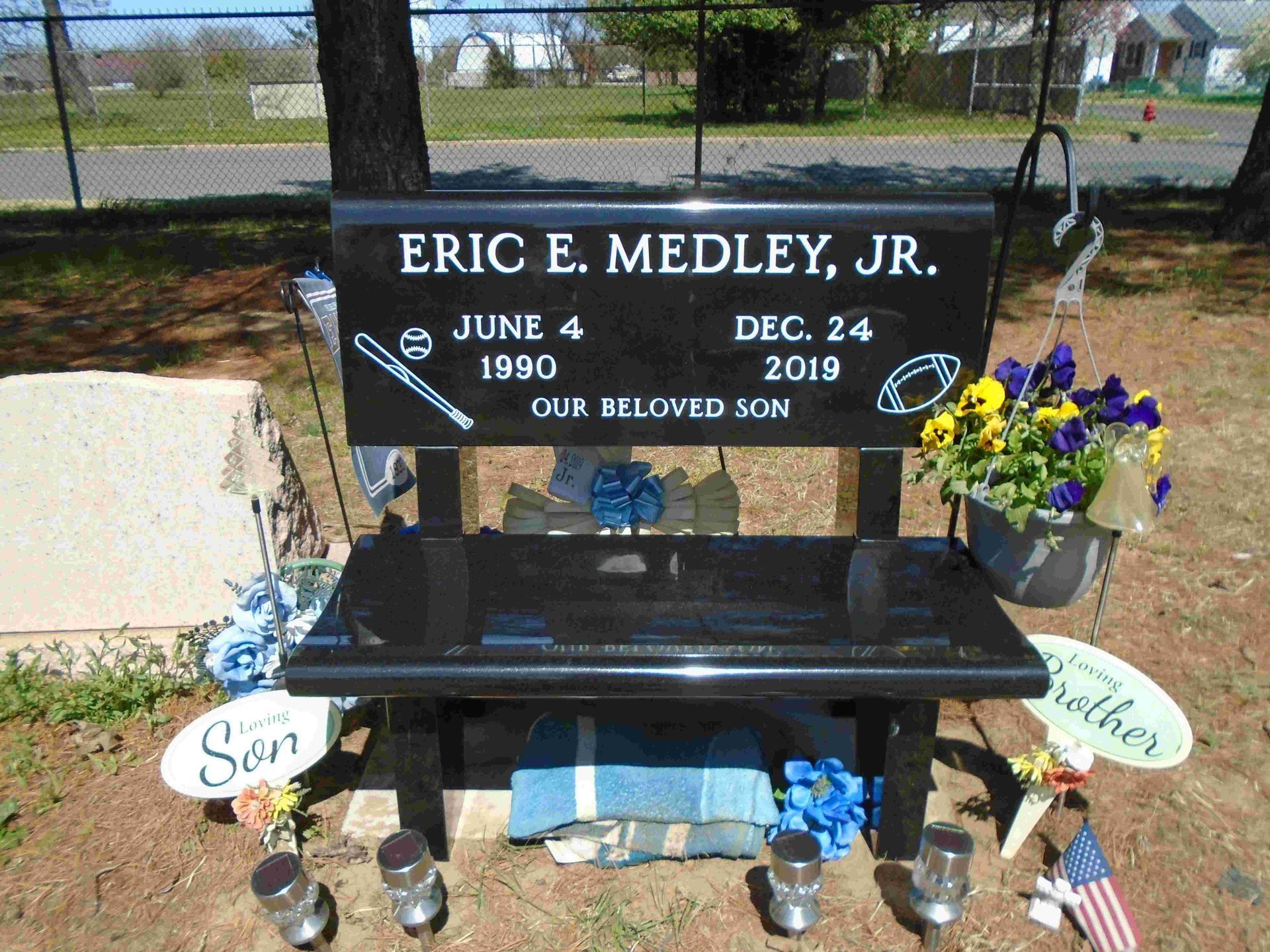 A black granite memorial bench for Eric E. Medley, Jr., engraved with birth and death dates, flowers, and decorations.