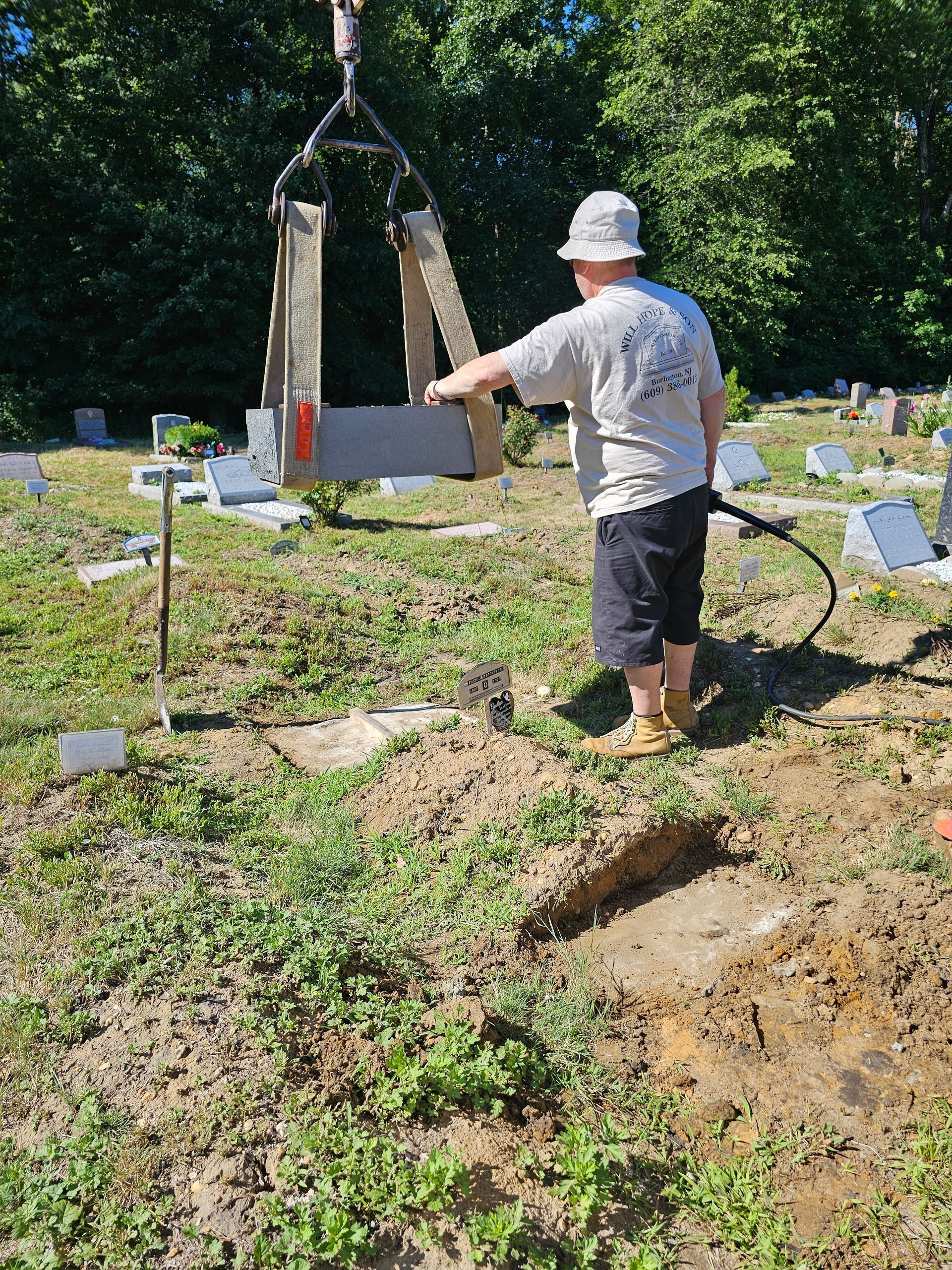 A person uses a mechanical hoist to lower a flat stone marker into a grave in a grassy cemetery.