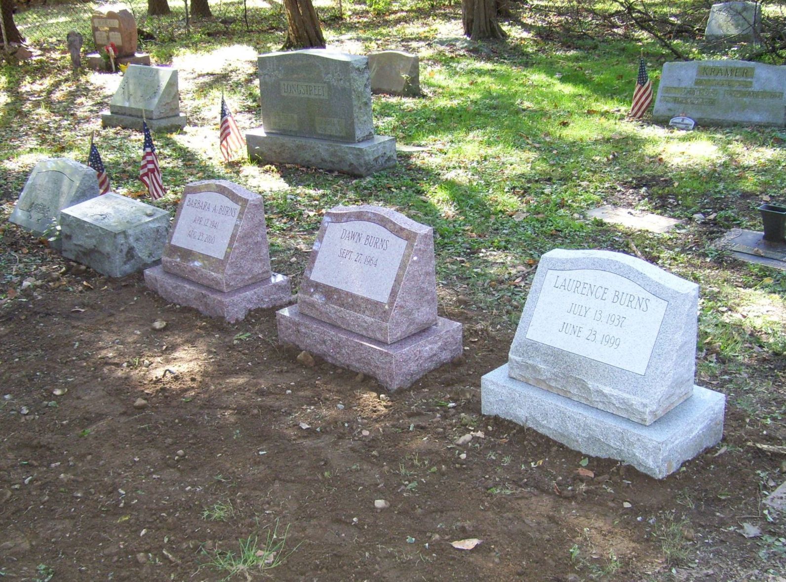 A sunny cemetery with rows of granite headstones, some with small American flags, on a patch of grass and dirt.