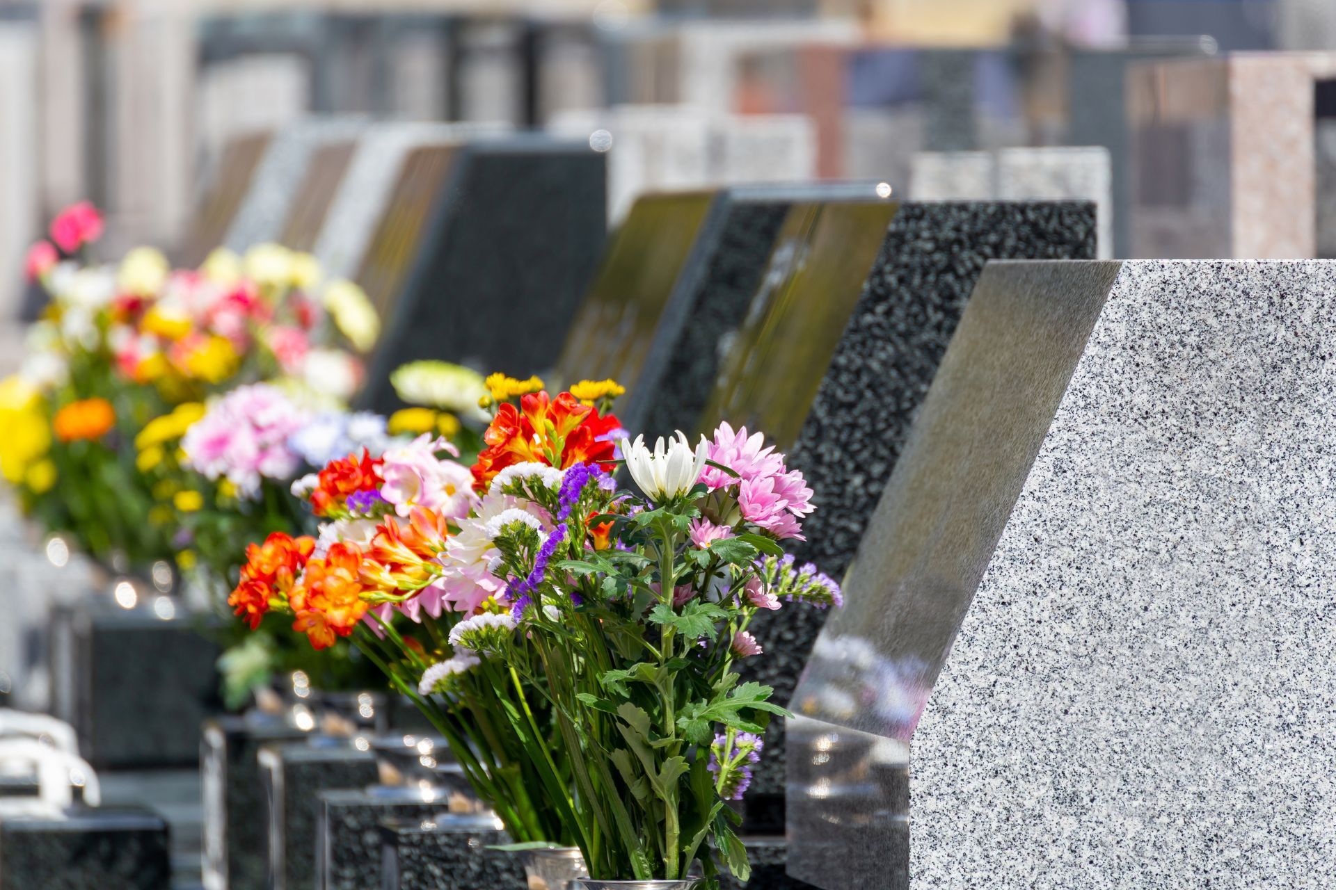 A row of granite gravestones in a cemetery, each adorned with colorful bouquets of fresh flowers.