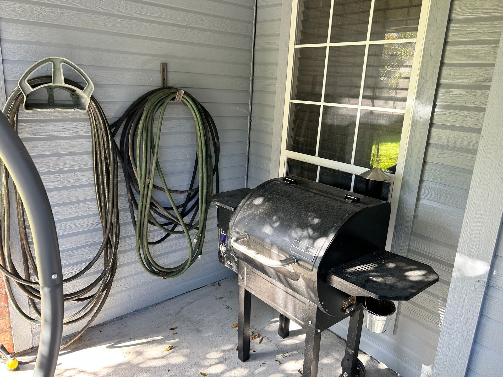Black grill next to a window on a porch, hoses hanging on the wall.